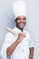 es un campeón en la cocina. alegre joven chef africano con uniforme blanco que lleva un rodillo en el hombro mientras se enfrenta a un fondo gris foto