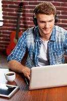 Doing creative work. Top view of handsome young man working on laptop and smiling while acoustic guitar laying in the background photo