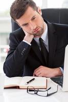 Tired businessman. Frustrated young man in formalwear looking away and touching his neck with hand while sitting at his working place photo