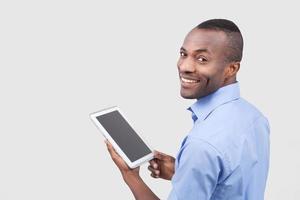 Working on digital tablet. Rear view of young African man working on digital tablet and looking over shoulder while standing isolated on grey photo