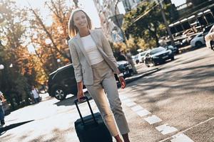 empresaria confiada. hermosa joven en traje tirando de equipaje y sonriendo mientras camina al aire libre foto