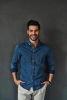 Irresistible man. Handsome young man smiling and keeping hands in pockets while standing against grey background photo