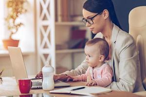 la mujer de negocios moderna puede manejar todo. vista lateral de una joven y hermosa mujer de negocios que usa una laptop mientras se sienta con una niña de rodillas en su lugar de trabajo foto