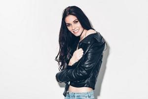 Flirty beauty. Beautiful young woman in unbuttoned leather jacket posing against grey background and smiling photo