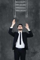 Helping ledder. Handsome young man in formalwear stretching out his hands and looking up while standing against chalkboard with drawing ledder on it photo