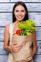 Health in her hands. Beautiful young woman in apron holding paper shopping bag full of fresh vegetables and smiling while standing in front of wooden background photo