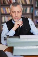 solo inspirado hombre mayor de pelo gris confiado en ropa formal sentado en la máquina de escribir y mirando a la cámara con estantería en el fondo foto