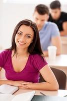 Happy student. Top view of beautiful female student sitting at the desk and smiling at camera while other students sitting in a row behind her photo