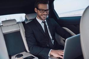 planificación de una nueva estrategia empresarial. un joven apuesto con traje completo que trabaja con una laptop y sonríe mientras está sentado en el auto foto