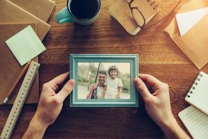 My son is my inspiration. Close-up top view of man holding photograph of himself and his son fishing over wooden desk with different chancellery stuff laying around photo