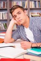 Tired student in library. Bored young man leaning his face on hand and looking at camera while sitting at the desk and in font of bookshelf