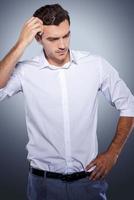 Lost in thoughts. Handsome young man in white shirt holding hand in hair and looking away while standing against grey background photo