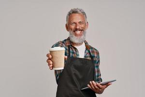 Senior man in apron smiling and giving you coffee while standing against gray background photo