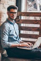 Working on his blog. Handsome young man working on laptop and looking at camera while sitting at windowsill photo