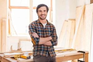 mi taller es mi mundo feliz joven carpintero con los brazos cruzados y apoyado en la mesa de madera con diversas herramientas de trabajo sobre ella foto