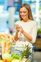 Inviting friends for dinner. Cheerful young woman holding mobile phone and smiling while standing in a food store photo