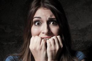 I am so scared Shocked young woman staring at camera while holding fingers in mouth while standing against dark background photo