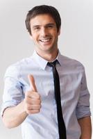 bien hecho joven feliz con camisa y corbata mostrando su pulgar hacia arriba y sonriendo mientras está de pie contra un fondo gris foto