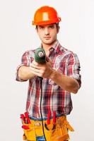 Freeze Handsome young handyman in hardhat stretching out drill and looking at camera while standing against grey background photo