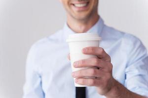 Fresh coffee for you Close-up of young man in shirt and tie stretching out coffee cup and smiling while standing against grey background photo