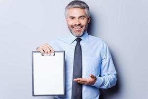 Your text here. Happy young man in formalwear holding clipboard and pointing it with smile while standing isolated on white background photo