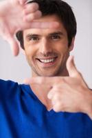 Focus on me Handsome young man in blue sweater gesturing finger frame and smiling while standing against grey background photo