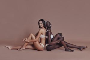 Feeling free to express sensuality. Two attractive mixed race women looking at camera while sitting back to back against brown background photo