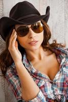 Western beauty. Beautiful young cowgirl adjusting her eyewear and looking at camera while standing against the wooden background photo