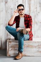 Stylish and confident. Handsome young man adjusting his sunglasses and looking at camera while sitting against the wooden wall photo