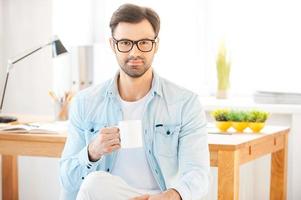 Cup of coffee for great ideas. Handsome young man in shirt and eyewear holding cup of coffee and looking at camera while sitting in front of his working place photo