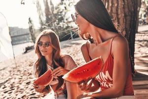Simply happy. Two attractive young women smiling and eating watermelon while sitting on the beach photo