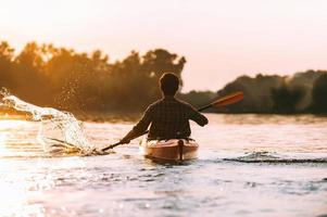 hombre en kayak. vista trasera del joven salpicando agua mientras navega en kayak en el río con la puesta de sol en el fondo foto