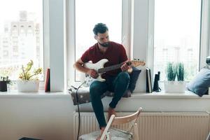 un joven apuesto con ropa informal tocando la guitarra mientras se sienta en el alféizar de la ventana foto