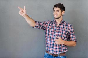 Pointing your advertisement. Happy young man pointing away and smiling while standing against grey background photo