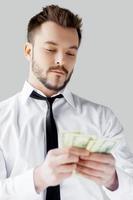 Young and wealthy. Confident young man in shirt and tie counting money and while standing against grey background photo