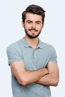 Candid smile. Handsome young man keeping arms crossed and looking at camera while standing against white background photo