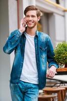 Ya estoy aquí, un joven alegre hablando por teléfono móvil y sonriendo a la cámara mientras estoy parado en un café en la acera foto