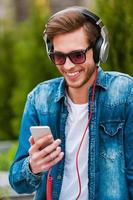 Me encanta esta canción, un joven feliz con auriculares sosteniendo un teléfono móvil y sonriendo mientras está sentado al aire libre foto