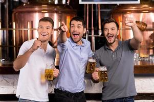 Goal Three happy men holding beer mugs and gesturing while watching TV in bar photo
