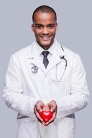 Your heart in right hands. Confident African doctor holding heart shape toy and smiling while standing against grey background photo