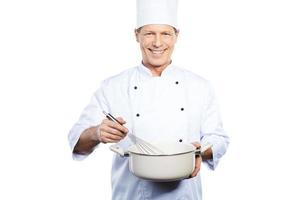 Chef at work. Cheerful mature chef in white uniform mixing something in casserole while standing against white background photo