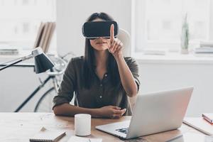 VR technologies. Confident young woman in virtual reality headset pointing in the air while sitting at her working place in office