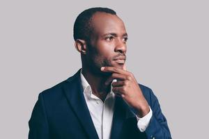 Lost in thoughts. Portrait of thoughtful young African man in smart casual jacket holding hand on chin and looking away while standing against grey background photo