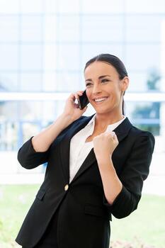 ganador de negocios. Hermosa mujer joven en ropa formal hablando por teléfono móvil y gesticulando mientras está de pie al aire libre foto
