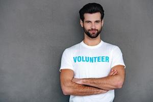 Ready for charity work. Confident young man in volunteer t-shirt keeping arms crossed and looking at camera with smile while standing against grey background photo