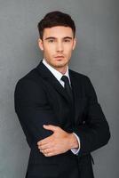 Young and successful. Confident young businessman keeping arms crossed and looking at camera while standing against grey background photo