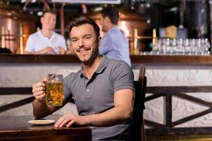 hombre en cervecería. joven alegre sosteniendo una jarra de cerveza y sonriendo mientras se sienta en el bar foto