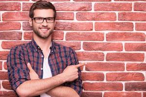 Confident handsome pointing your advertising. Handsome young man keeping arms crossed and pointing at copy space while standing against brick wall photo