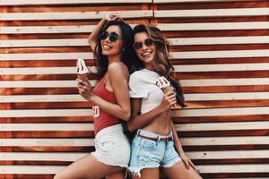 sentirse libre y feliz. dos atractivas mujeres jóvenes sonriendo y sosteniendo helado mientras están de pie contra la pared de madera al aire libre foto