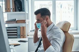 joven empresario de confianza con camisa y corbata poniéndose una máscara protectora mientras está sentado en la oficina foto